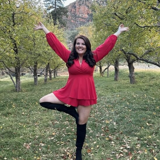 A person in a red dress and black boots stands on one leg in a tree pose, outdoors in a grassy area with trees in the background.