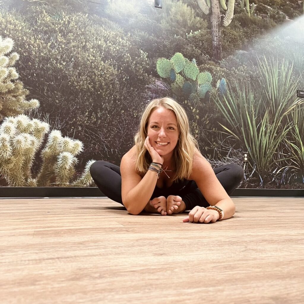 Person in a yoga pose on a wooden floor, smiling with one hand on their cheek. Background features a desert-themed wall mural with cacti and greenery.