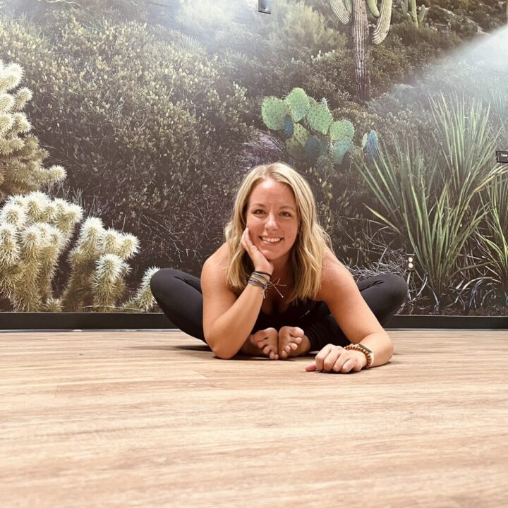 Person in a yoga pose on a wooden floor, smiling with one hand on their cheek. Background features a desert-themed wall mural with cacti and greenery.