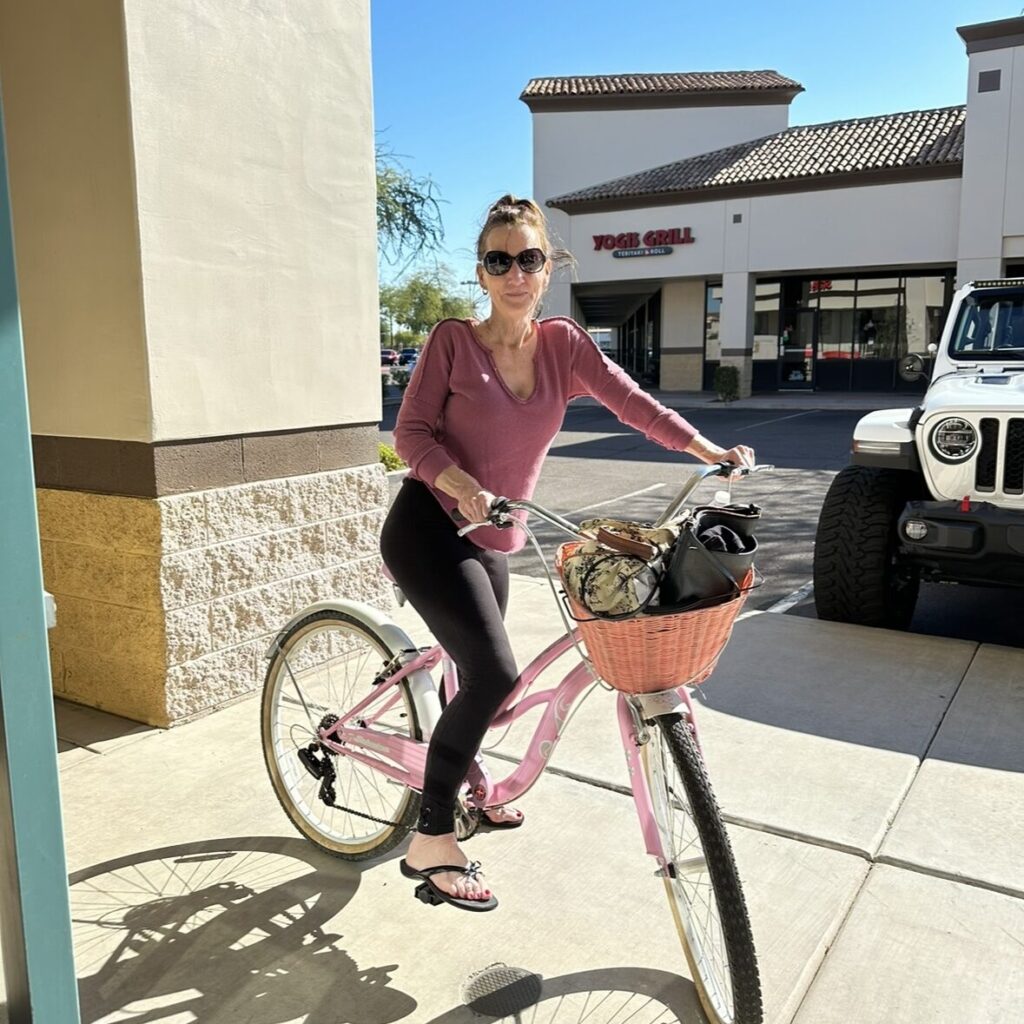 Person in sunglasses and pink top stands with a pink bicycle outside a shopping area on a sunny day.