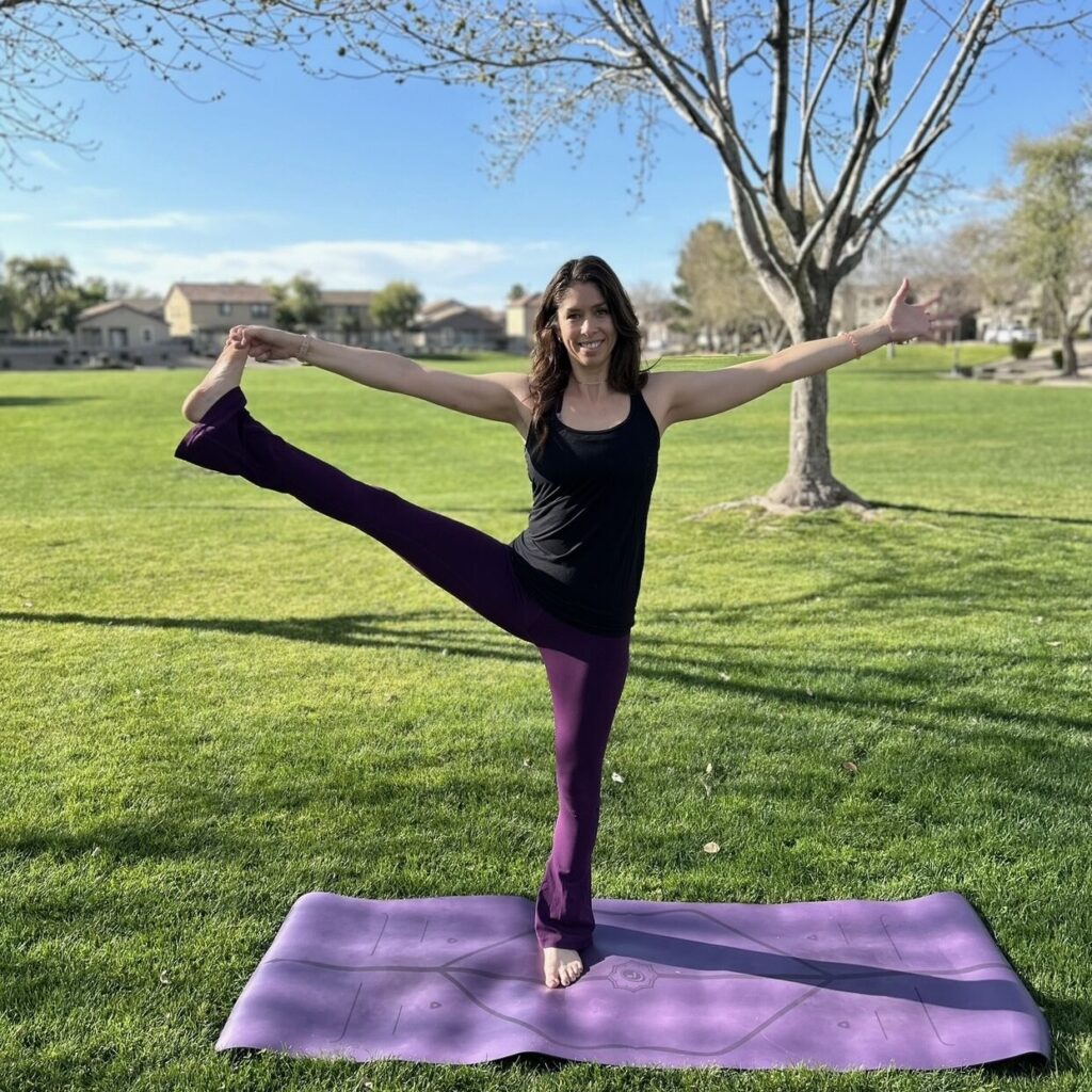 A person practices yoga on a purple mat in a park, balancing on one leg with the other leg extended. Trees and houses are visible in the background under a clear blue sky.