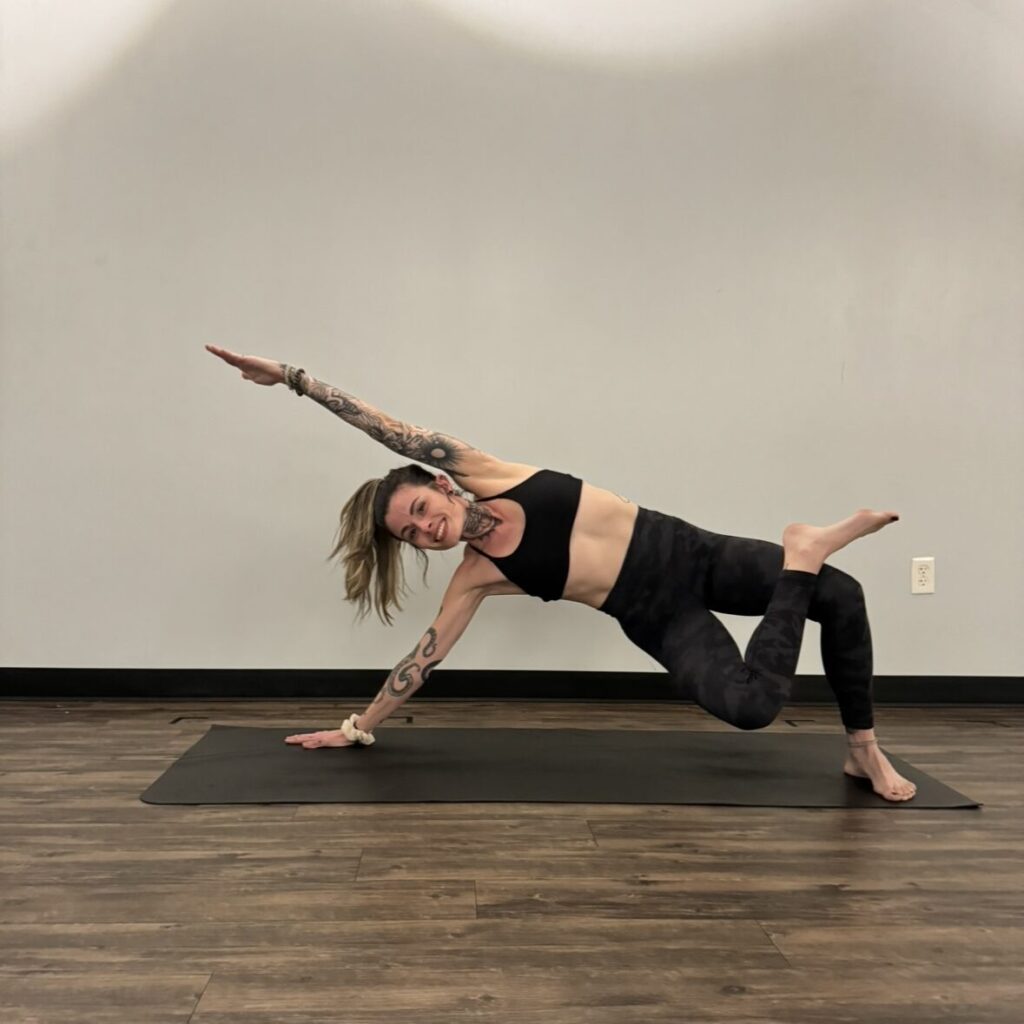Person performing a side plank variation on a yoga mat in a room with wooden flooring and a plain wall.