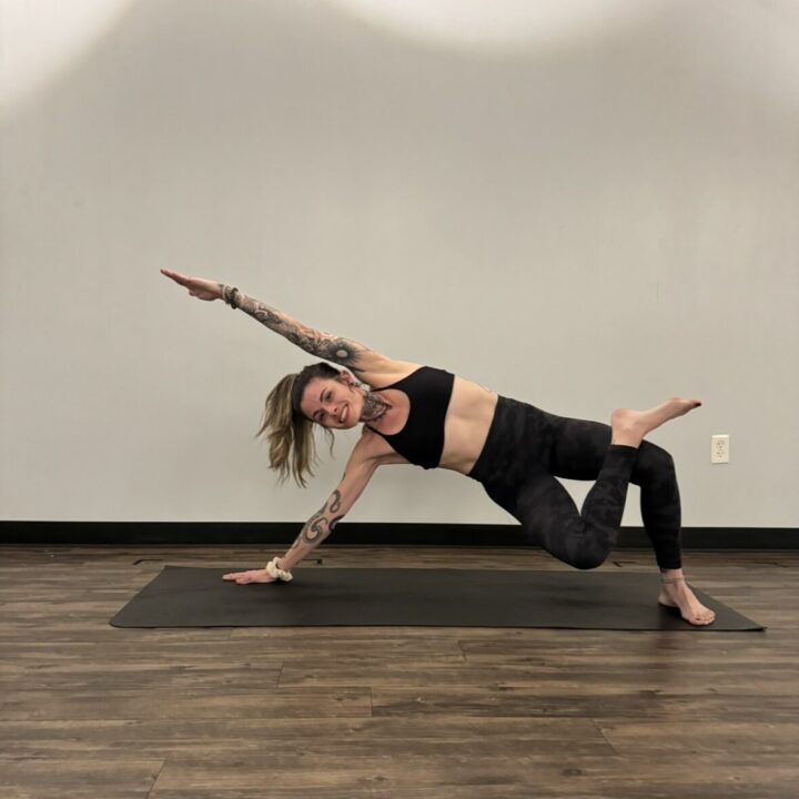 Person performing a side plank variation on a yoga mat in a room with wooden flooring and a plain wall.