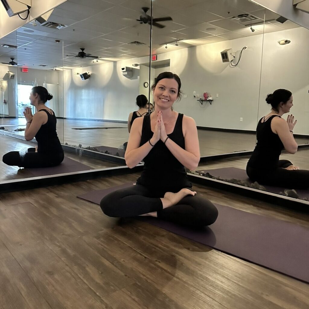 Person sitting cross-legged on a yoga mat in front of a mirrored wall, hands in a prayer position, wearing black workout clothes in a studio.