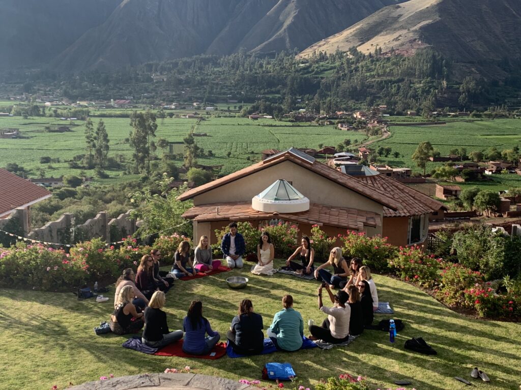 A group of people sit in a circle on a grassy hill, surrounded by mountains, with a building in the background.