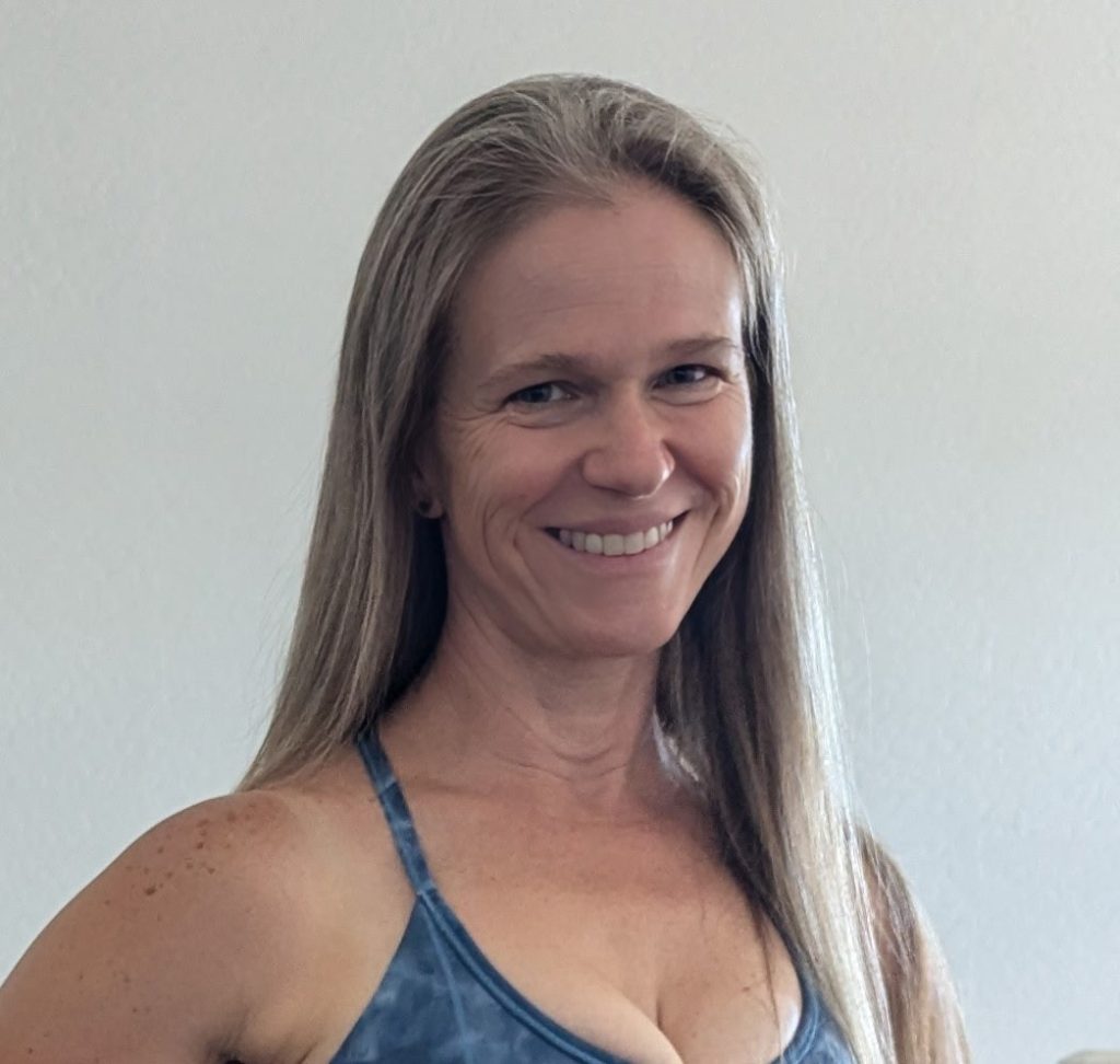 A woman with long straight hair, wearing a blue sleeveless top, stands smiling in front of a plain light-colored wall.