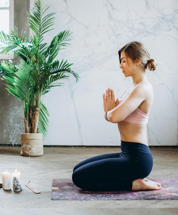 A person sitting in a meditative pose on a yoga mat, with hands in prayer position. There are lit candles and a potted plant nearby, against a marble wall background.