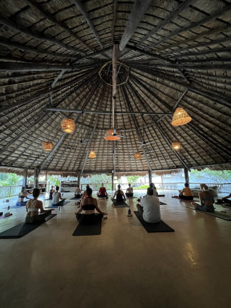 People are seated on yoga mats under a large, open, wood-framed hut with a thatched roof. Hanging lights illuminate the dim space.