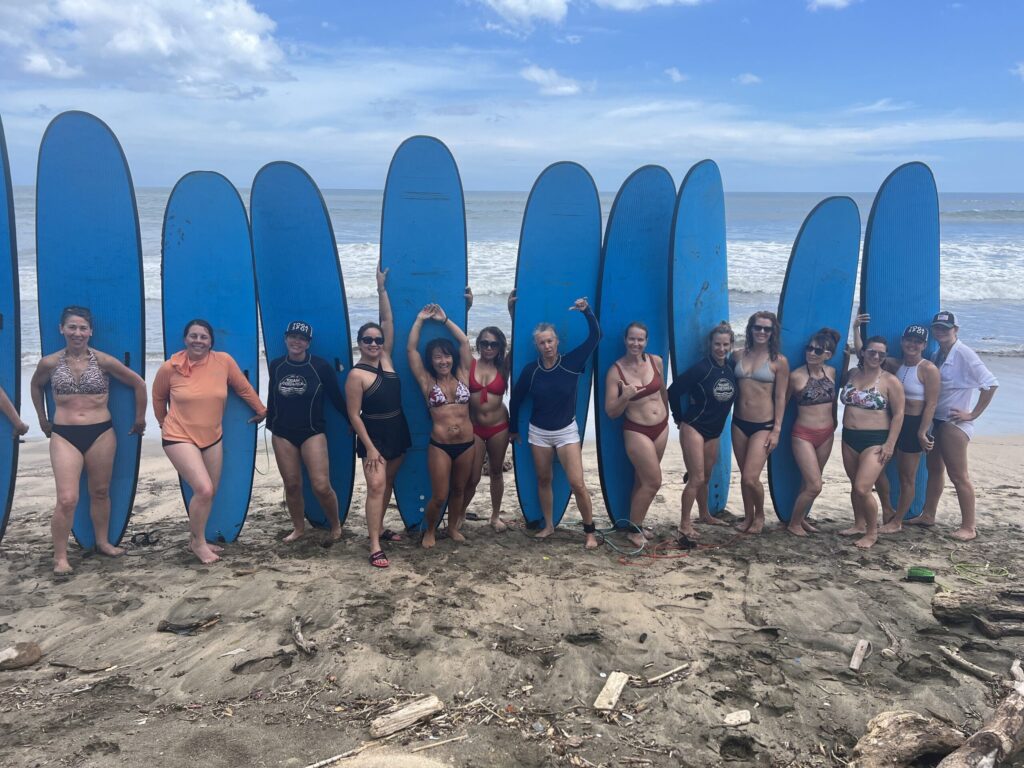 A group of people in swimwear stand in front of blue surfboards arranged vertically on a sandy beach. The ocean and cloudy sky are visible in the background.