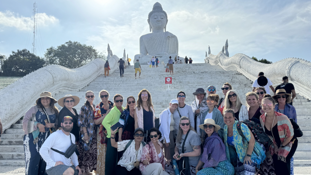 A group of people posing together on stone steps leading up to a large statue of Buddha under a partly cloudy sky.