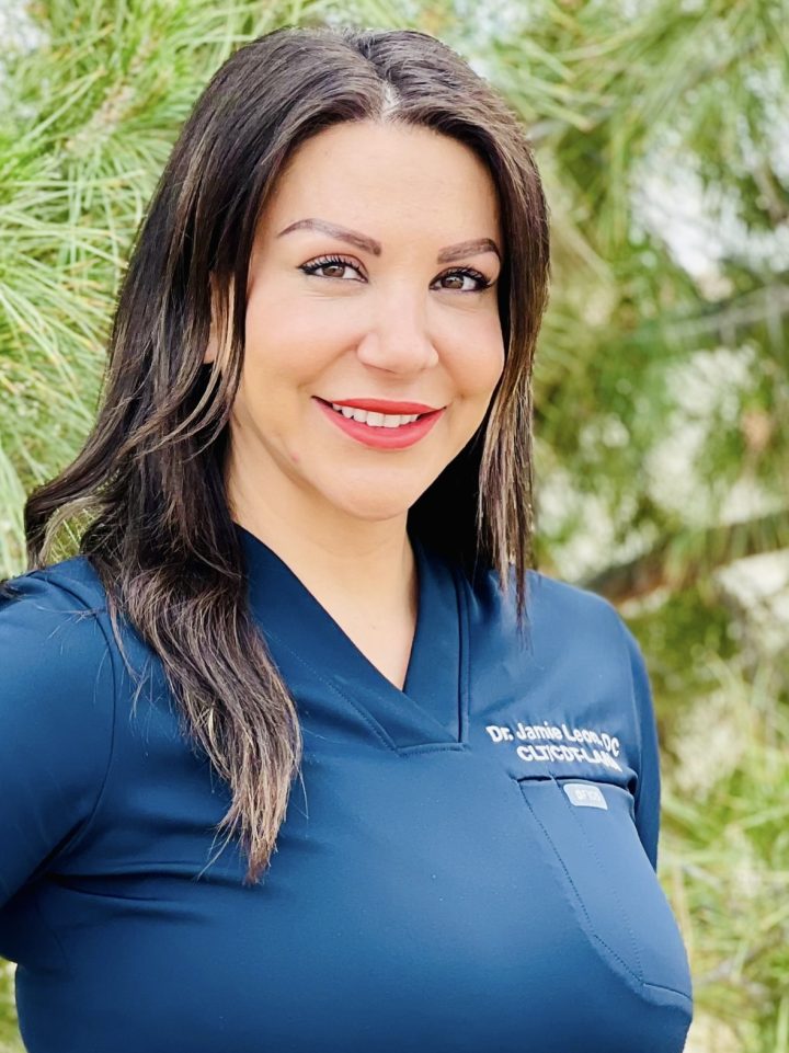 A woman in a navy blue medical scrub top stands outdoors in front of green pine trees, smiling at the camera.