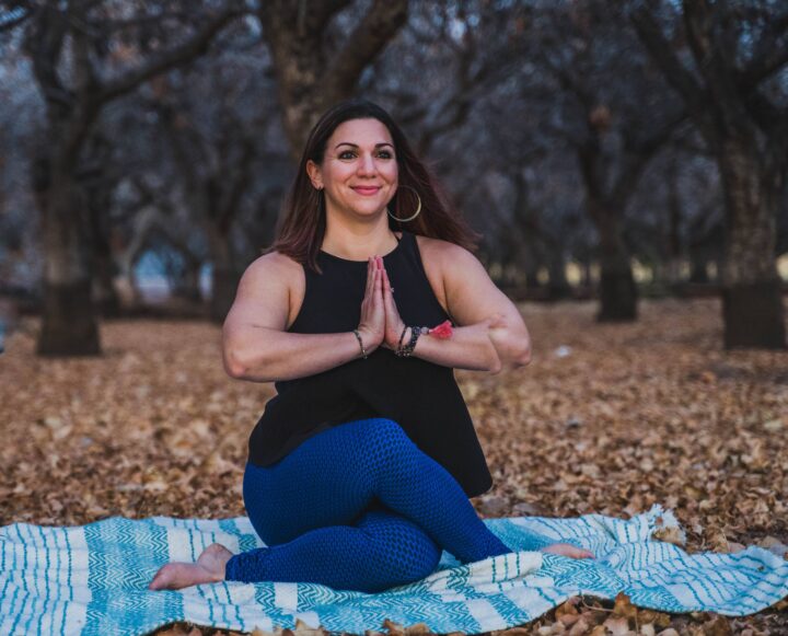A person sitting cross-legged in a meditative pose on a blanket in a park surrounded by autumn leaves and trees.