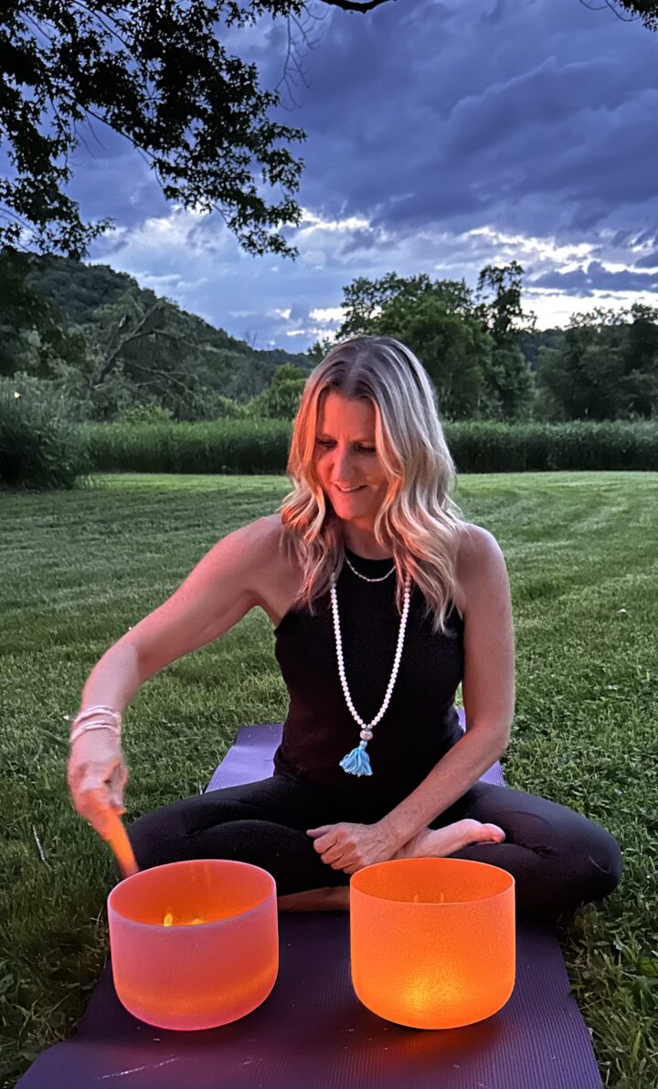 A woman sits cross-legged on a mat outdoors, playing two orange crystal singing bowls with a mallet. She is wearing a black outfit and a beaded necklace. Trees and a cloudy sky are in the background.