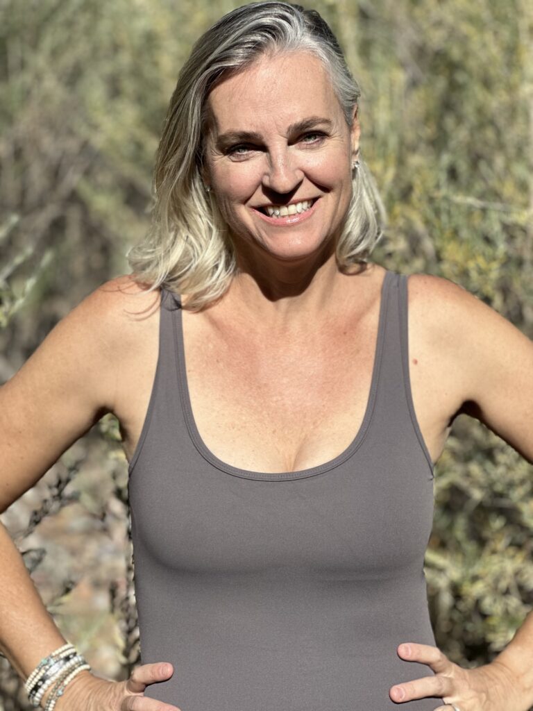 Person smiling outdoors, wearing a gray tank top, with arms on hips, surrounded by blurred greenery.