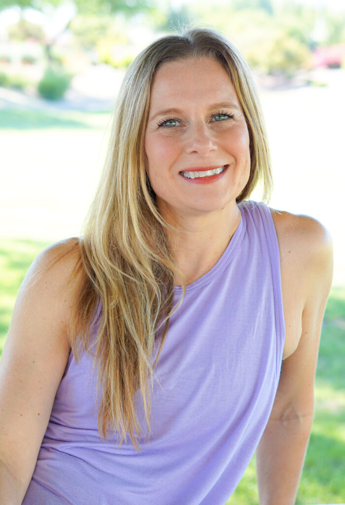 Woman with long blonde hair wearing a sleeveless purple top, smiling outdoors with a blurred natural background.