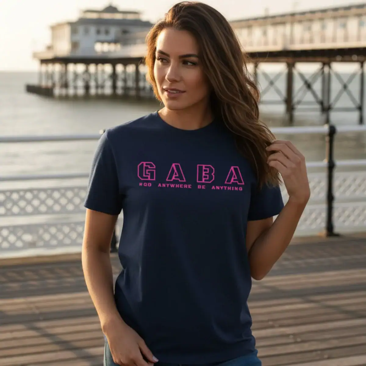 Woman wearing navy GABA Clothing T-shirt with logo, seaside background.