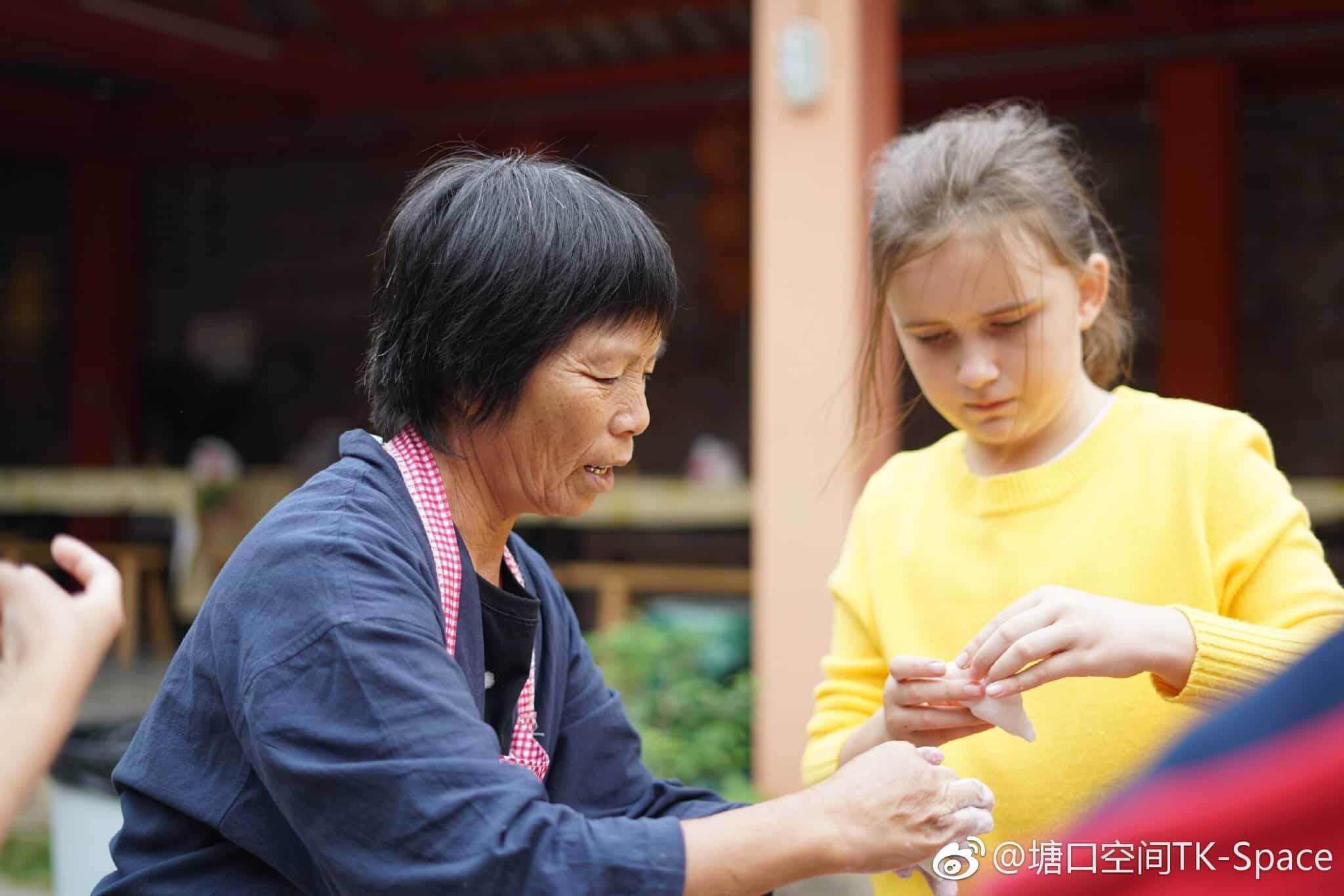 An elderly woman and a young girl working together on a hands-on activity at the Tangkou Community Project in Kaiping Tangkou, with traditional architecture in the background.