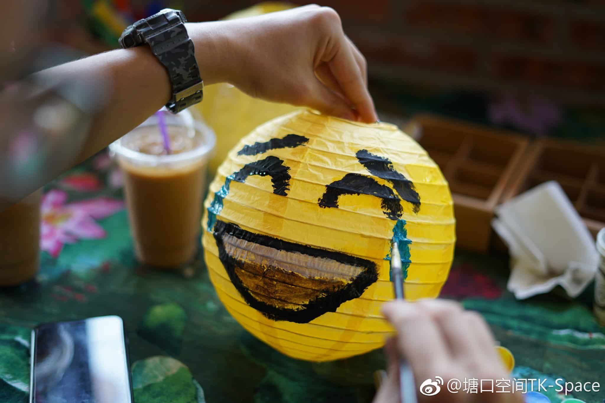 Hands painting a yellow paper lantern with a laughing emoji design during a community event at the Tangkou Community Project in Kaiping Tangkou, with iced coffee drinks on the table.