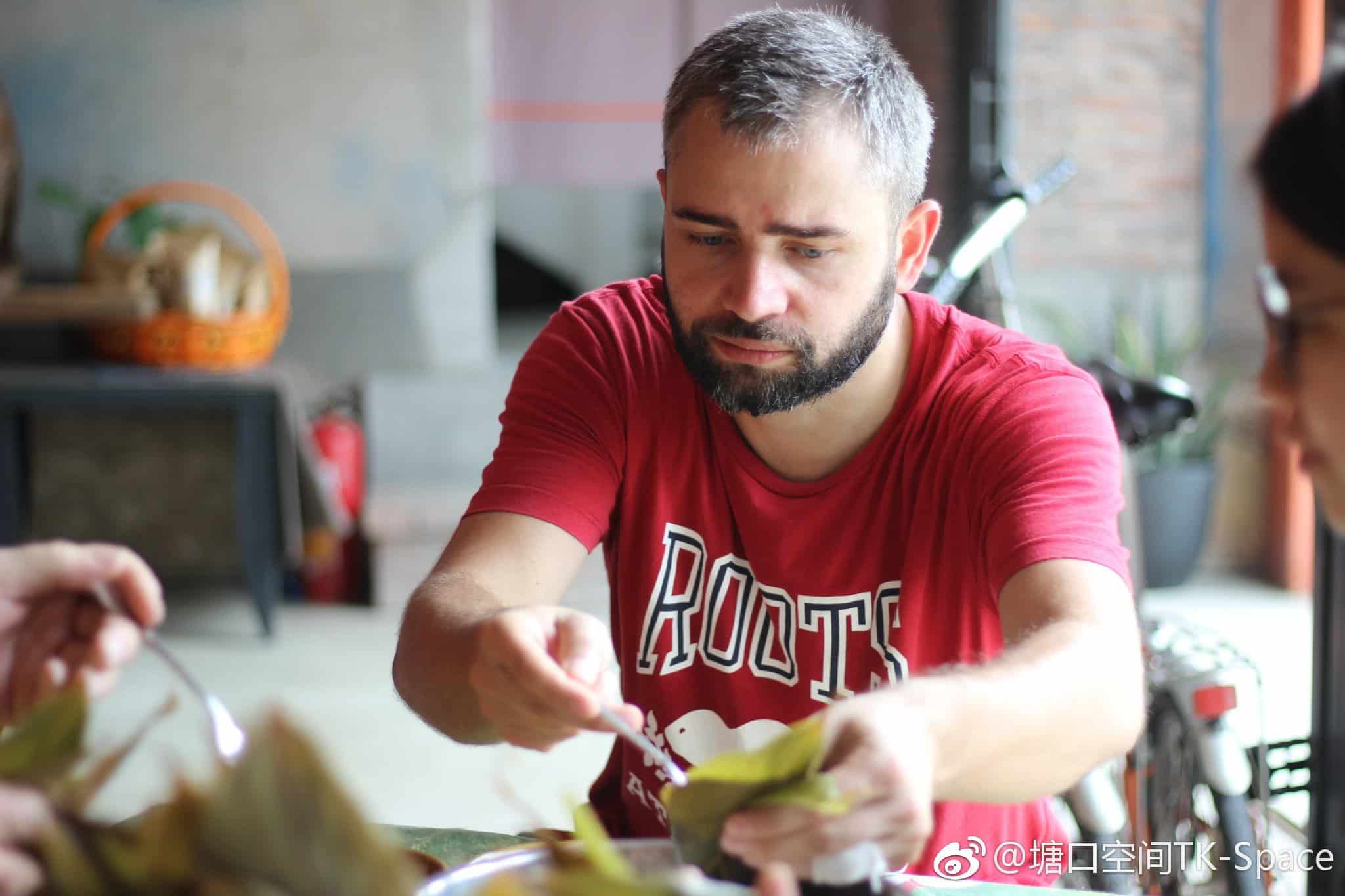 A man making zongzi with bamboo leaves at the Kaiping Tangkou Community Project, showcasing cultural heritage in a rustic indoor setting.