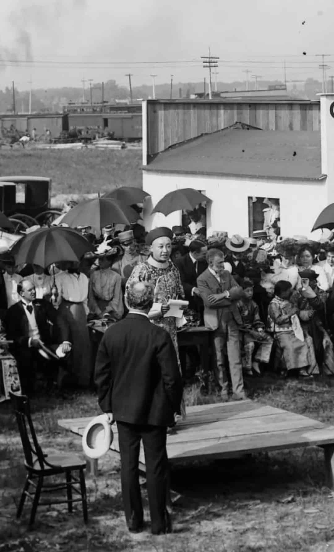 A black-and-white photograph of a large crowd gathered outdoors at an event, with individuals dressed in period clothing. The central figure is wearing traditional Chinese attire, standing on a platform surrounded by attendees holding umbrellas. The setting includes early 20th-century vehicles and structures in the background.