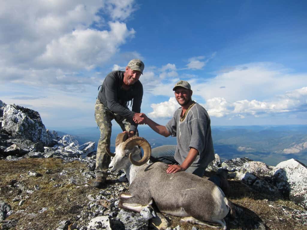 Two men shaking hands over a mountain goat on a rocky summit.