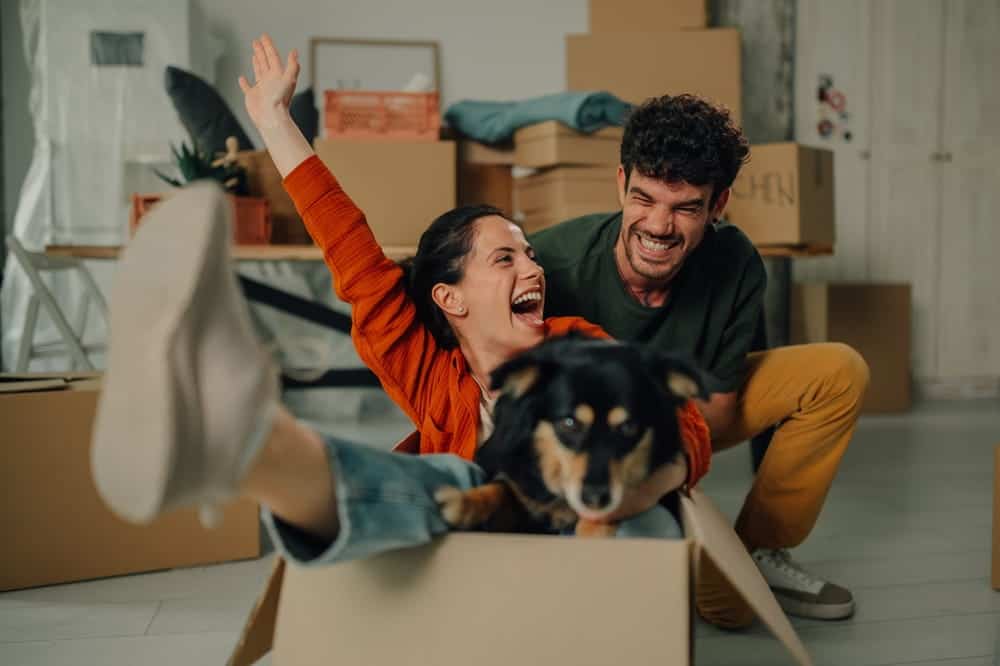 Alt text: Couple celebrating in their new home with their dog while surrounded by moving boxes.