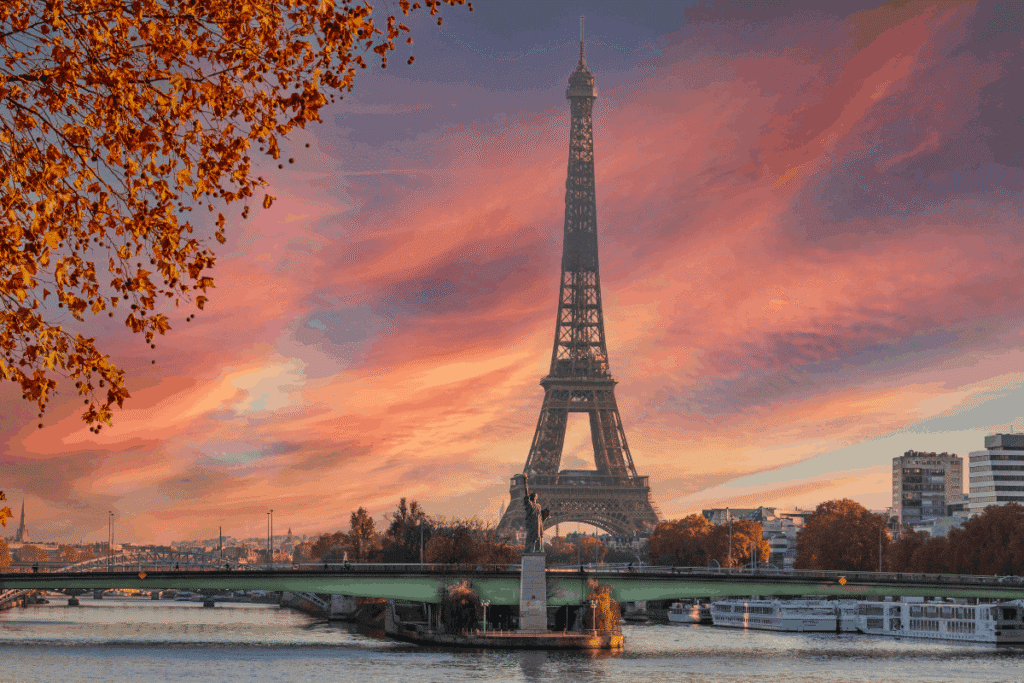 Eiffel Tower silhouetted against a vivid pink and orange sunset sky, with a bridge over the Seine River and autumn foliage in the foreground.