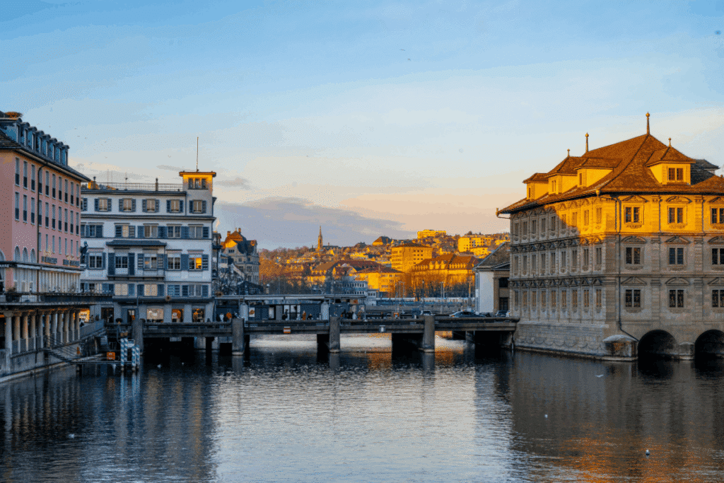 Zurich, Switzerland cityscape at sunset, showing historic buildings along the Limmat River with a bridge and reflections on the water.