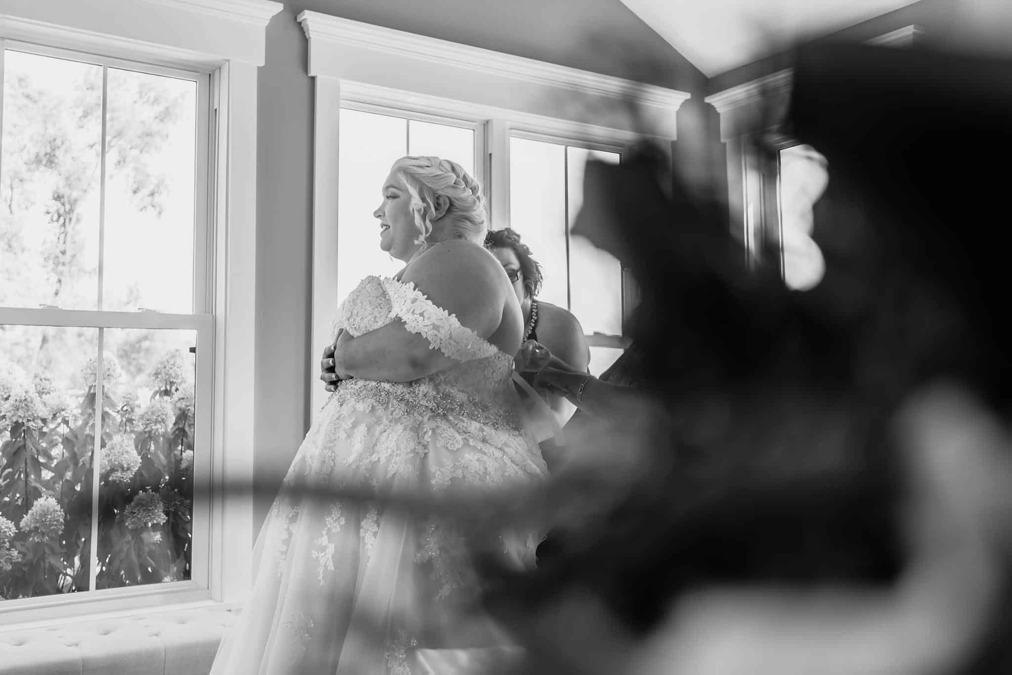 Black and white photo of bride standing still while being laced into dress during intimate fall wedding at the Lodge at River Valley in Yorktown, Indiana