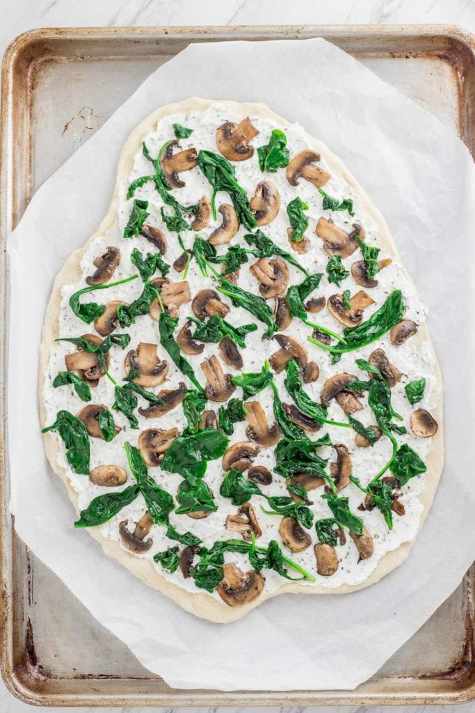 Whole white pizza on the baking sheet, ricotta cheese spread and sautéed mushroom and spinach on top of the pizza