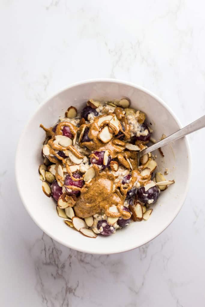 overnight oats in a white bowl with red grapes, almond slices, and almond butter drizzled on top