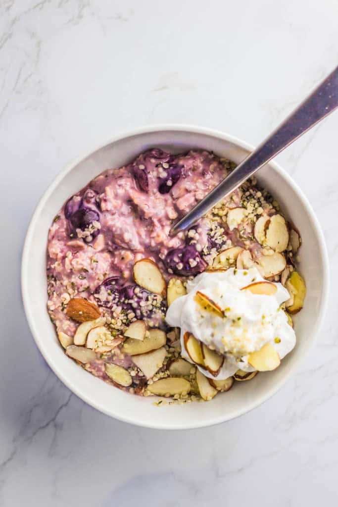Frozen cherry overnight oats in a white bowl with cashew yogurt, coconut whip cream, sliced almonds, and hemp seeds on top