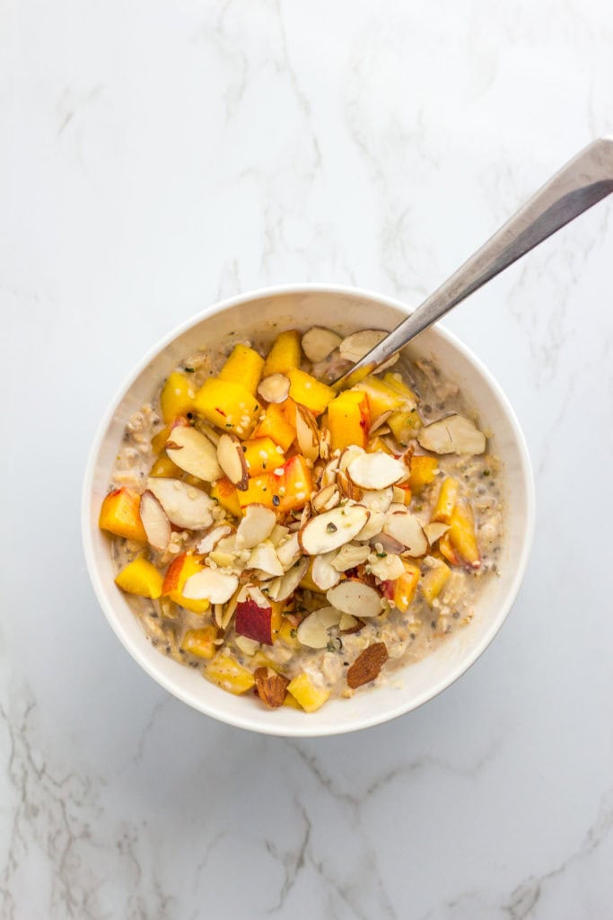 Overnight oats in a white bowl with diced peach, hemp seeds, and sliced almond on top