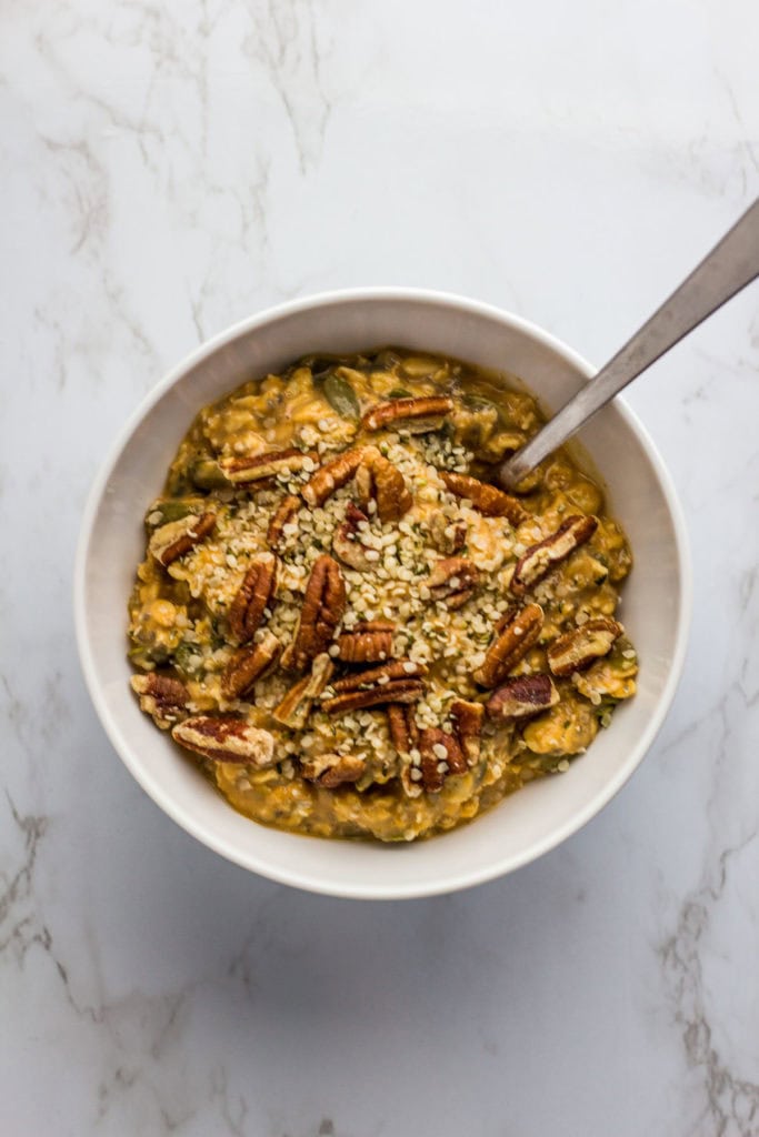 overnight oats in a white bowl with pumpkin puree, pumpkin spice, and pumpkin seeds. Sprinkled of pecan halves with hemp seeds