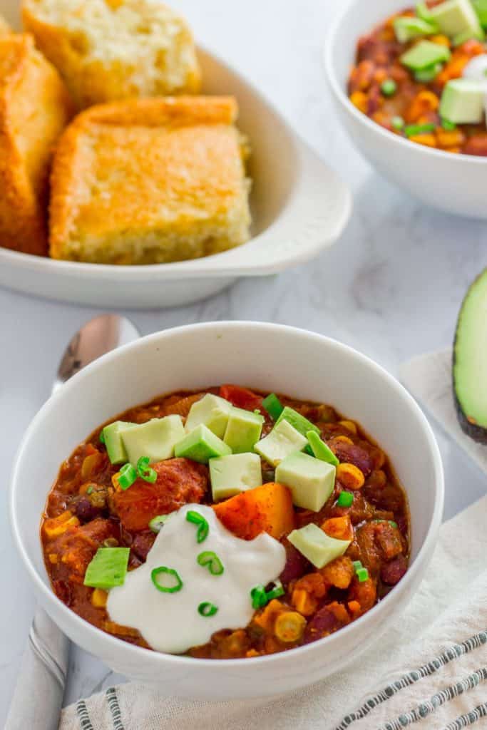 Bowl of vegan butternut squash chili topped with avocado and cashew cream, served with cornbread on the side.