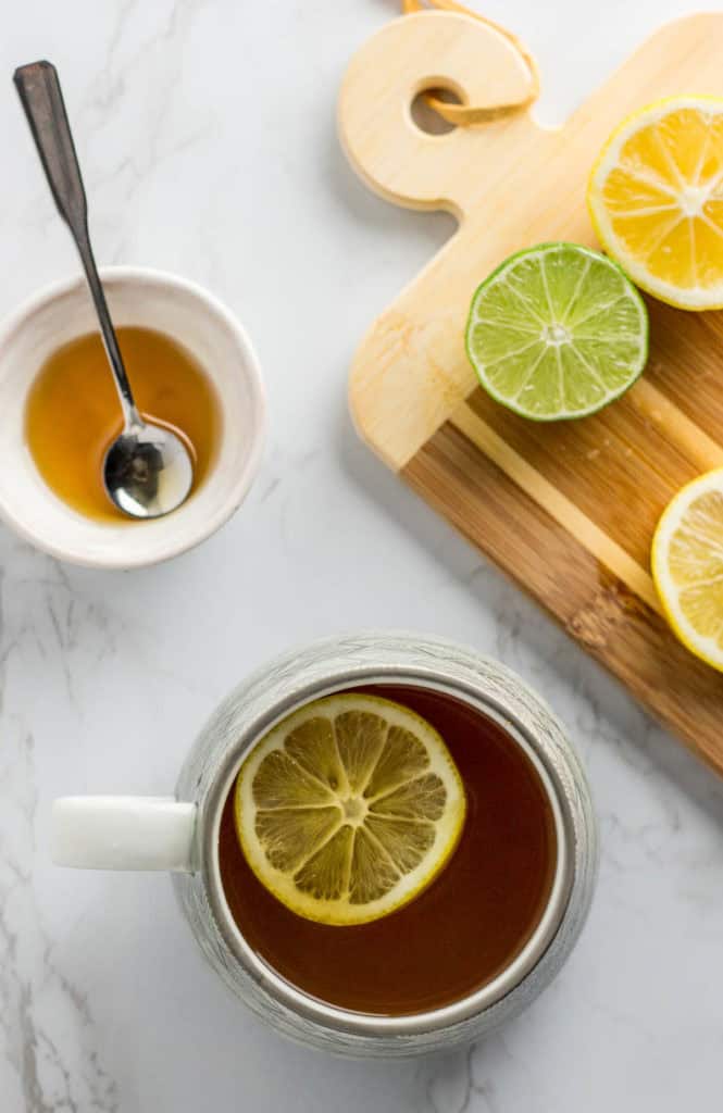 White mug filled with immune boosting tea topped with a lemon slice, next to a small bowl of honey and sliced lemon and lime on a cutting board.