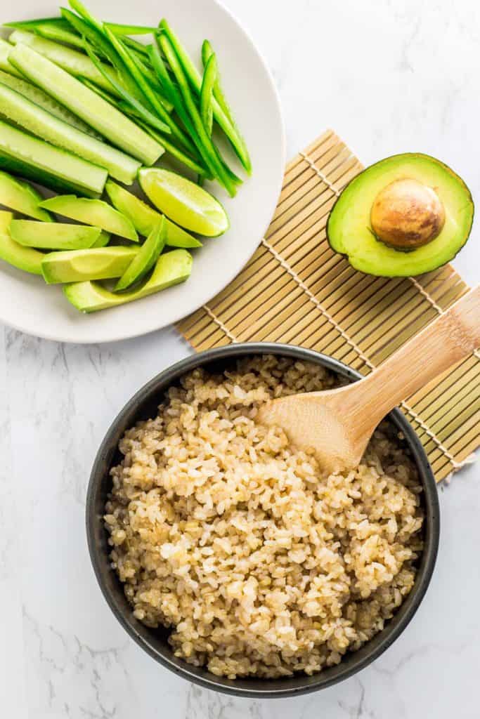 Over head shot of cucumber, avocado, and jalapeno slices on a white plate and seasoned sushi rice in a large black bowl.
