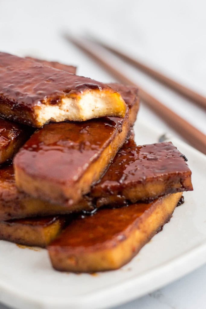 Close-up of oven-baked hoisin tofu slices stacked on a plate, glossy and saucy with a caramelized hoisin glaze
