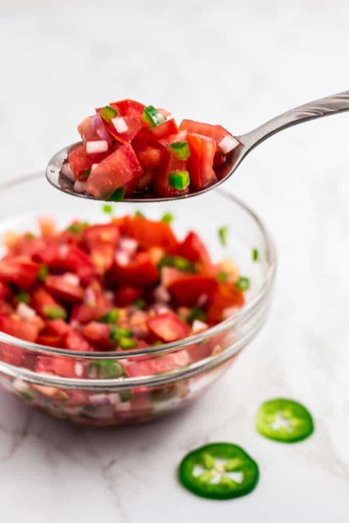 Spoonful of pico de gallo with tomato, onion, and jalapeño held over a glass bowl.