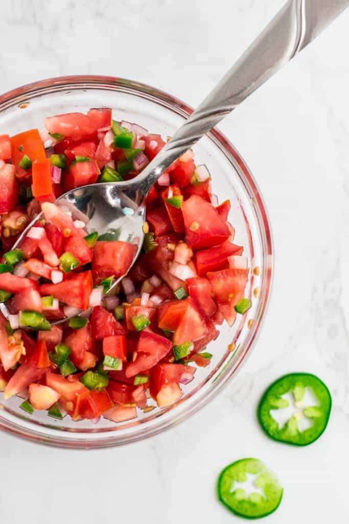 Easy pico de gallo recipe without cilantro in a glass bowl with spoon, made with tomato, onion, jalapeño, and red wine vinegar.