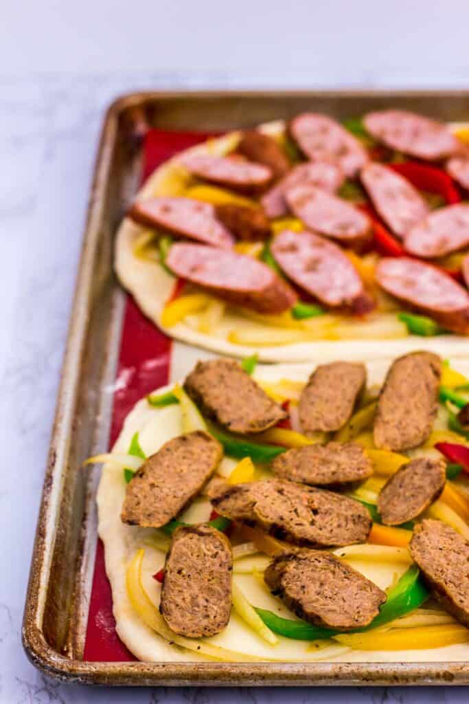 two pizza crusts topped with provolone cheese and sauteed onion and pepper. One pizza with chicken sausage and the other with vegan sausage