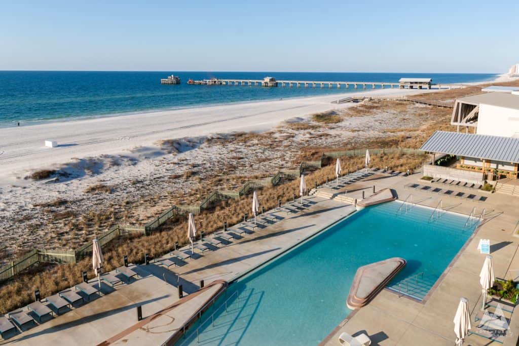 An aerial view of a swimming pool area at a Gulf Shores hotel, overlooking a pristine beach and the Gulf of Mexico. Lounge chairs with closed umbrellas line the pool deck, which is adjacent to a natural landscape of dunes and sea grass. In the distance, a long pier extends out into the ocean. The beach stretches along the horizon, with calm blue water under a clear sky.
