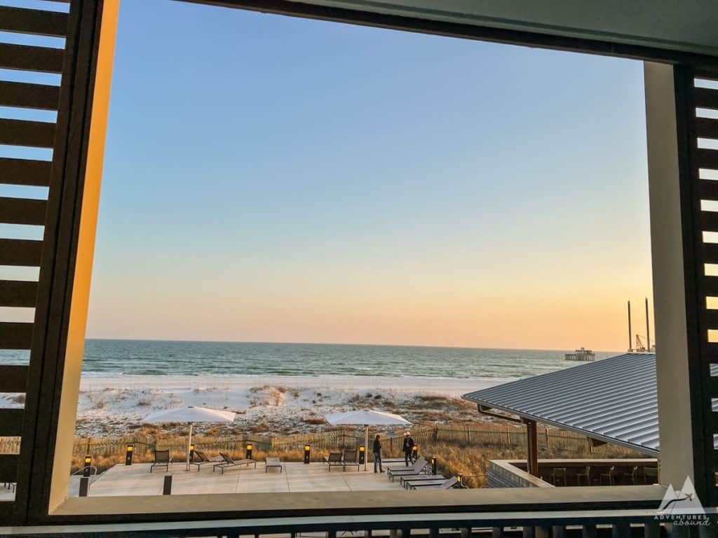 The image captures a beautiful sunset view of the Gulf of Mexico from a framed balcony perspective. The beach and ocean stretch across the horizon, with soft pastel hues in the sky. Below, lounge chairs and umbrellas are set up on a patio near the sandy shore, and a metal-roofed building is visible to the right. Dunes and sea grass border the beach, adding to the coastal atmosphere. The "Adventures Abound" logo is seen in the lower right corner.