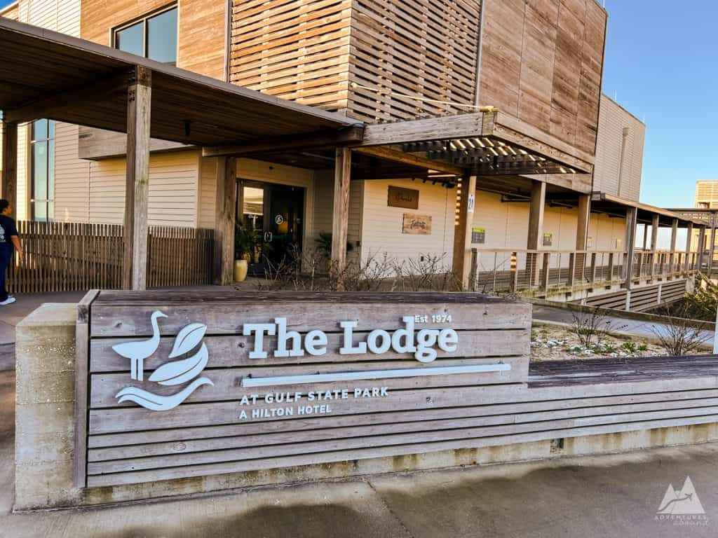 The entrance to The Lodge at Gulf State Park, a Hilton Hotel. The exterior features natural wood accents with slatted designs and a covered walkway leading to the main doors. A large wooden sign in the foreground displays the hotel's name and logo, which includes the image of a bird and leaves, along with the text "The Lodge, Est 1974, At Gulf State Park, A Hilton Hotel."