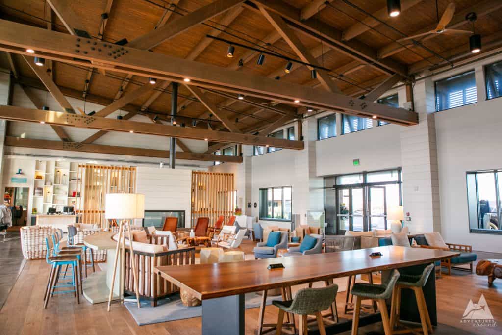 View of interior of a hotel lobby at The Lodge, featuring high ceilings with exposed wooden beams. The seating area includes a variety of chairs and couches in neutral and blue tones, arranged around low tables, creating a cozy, welcoming atmosphere. A long wooden communal table with stools is also visible, along with modern décor, large windows, and warm lighting. In the background, a small retail section can be seen beside the main check-in desk which sits behind a wall of spaced wooden slats.