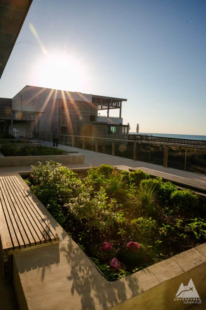 The image shows a building The Lodge hotel at Gulf State Park during sunset. The sun shines brightly over the structure, casting lens flares, while in the foreground, an edible garden with a variety of green plants and herbs is neatly arranged along concrete pathways. In the background, a boardwalk extends toward the ocean, where beach umbrellas are visible by the water. The lower right corner includes a logo for "Adventures Abound."