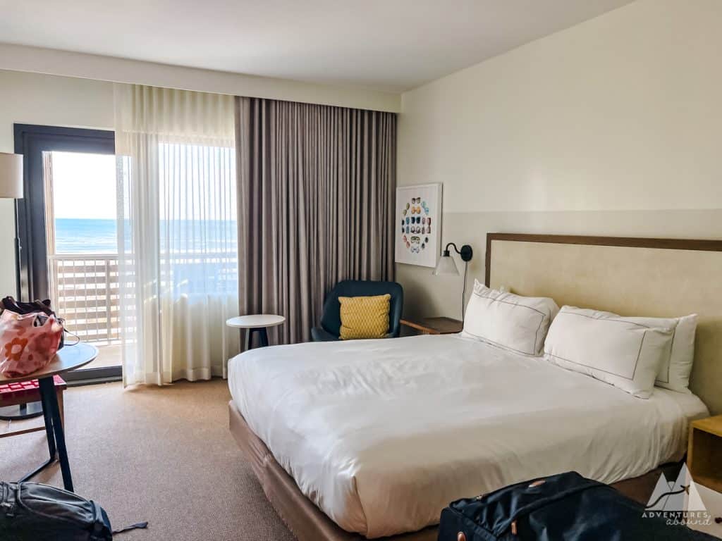 
The image shows a well-lit hotel room at The Lodge at Gulf State Park. The room features a king-sized bed with white linens, a small seating area with a dark chair and a yellow cushion, and a round side table. A sliding glass door opens to a balcony with ocean views, letting in natural light through sheer curtains. Luggage and bags are placed near a table, and a framed art piece with colorful sunglasses decorates the wall. The "Adventures Abound" logo is visible in the bottom right corner.