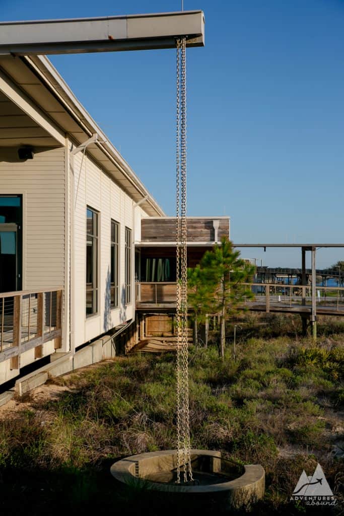 The image shows a sustainable rainwater chain hanging from the roof of a modern building at The Lodge in Gulf State Park. The chain leads down into a circular stone basin, surrounded by native grasses and plants, emphasizing eco-friendly design. This system allows rainwater to be absorbed by the wetland ecocystem on the property. A wooden walkway and building with large windows are visible in the background under a clear blue sky