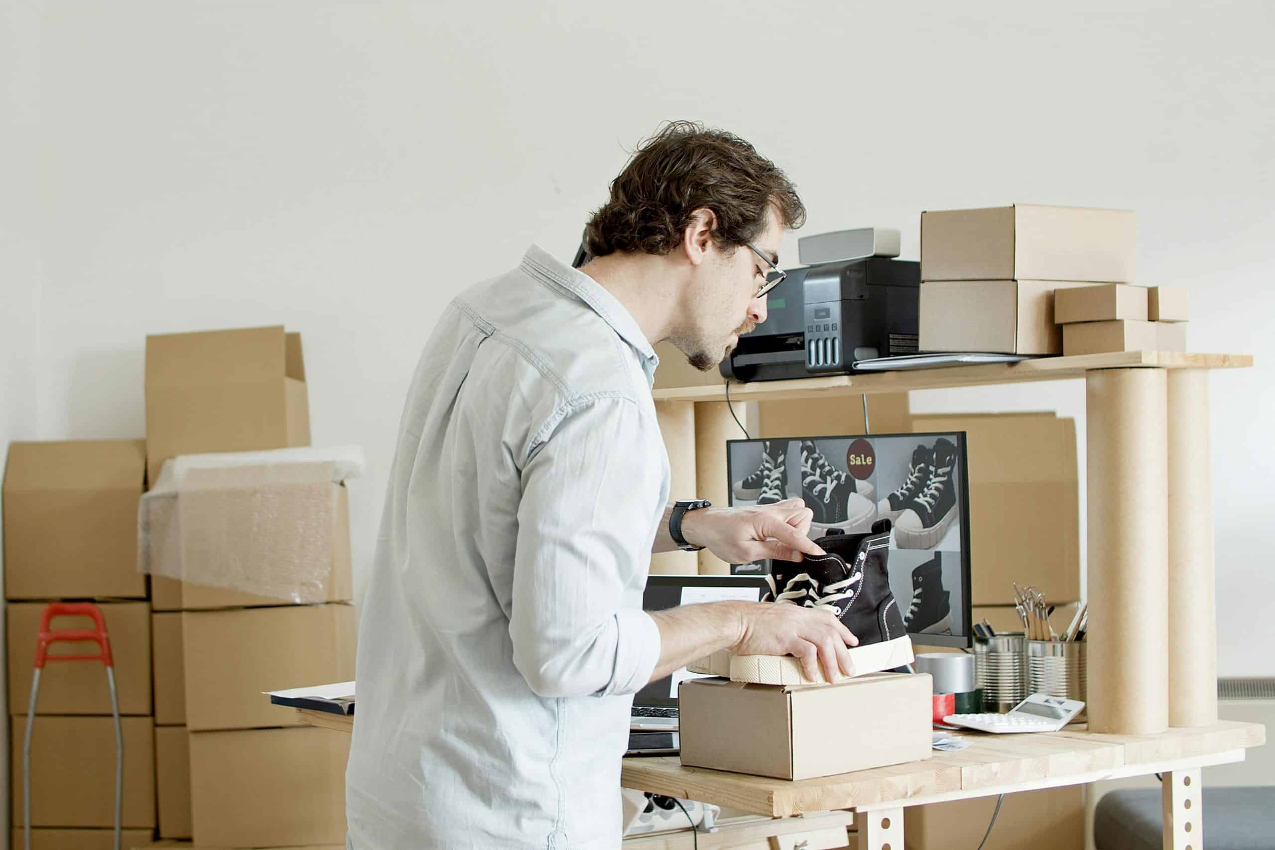 A man packing black sneakers into a box in a workspace filled with cardboard packages.