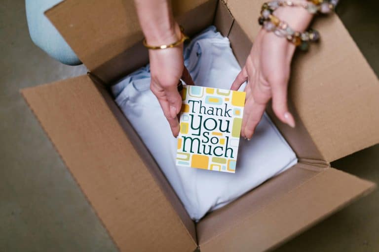 A person packing a package containing clothing with a "Thank You So Much" card on top.